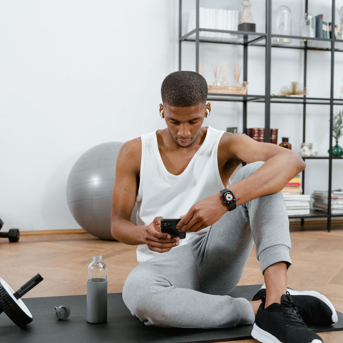 A man sitting on his exercise mat at home, checking his phone, with a water bottle and an ab wheel beside him.