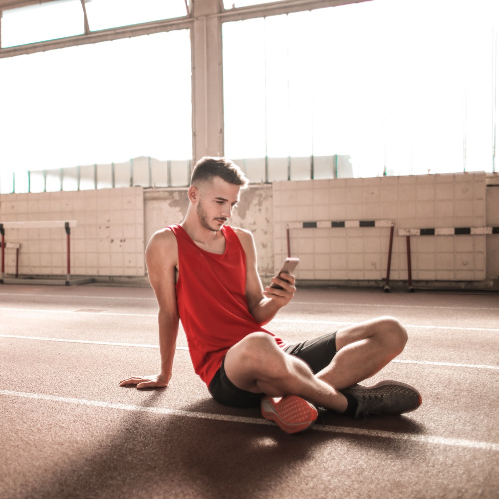 A man in a red vest sitting on an indoor running track, smiling at his phone.
